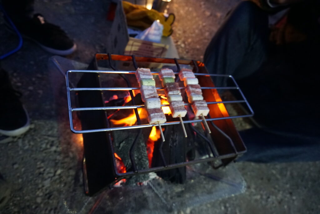 焚き火の上に置いた網で焼いている豚串